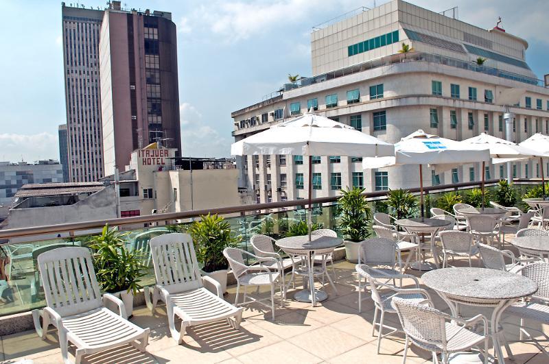 Hotel Atlântico Business Centro Río de Janeiro Exterior foto The photo shows a rooftop area with several white lounge chairs and circular tables. There are umbrellas providing shade over some of the seating. In the background, there are tall buildings, including a modern skyscraper with a distinct architectura